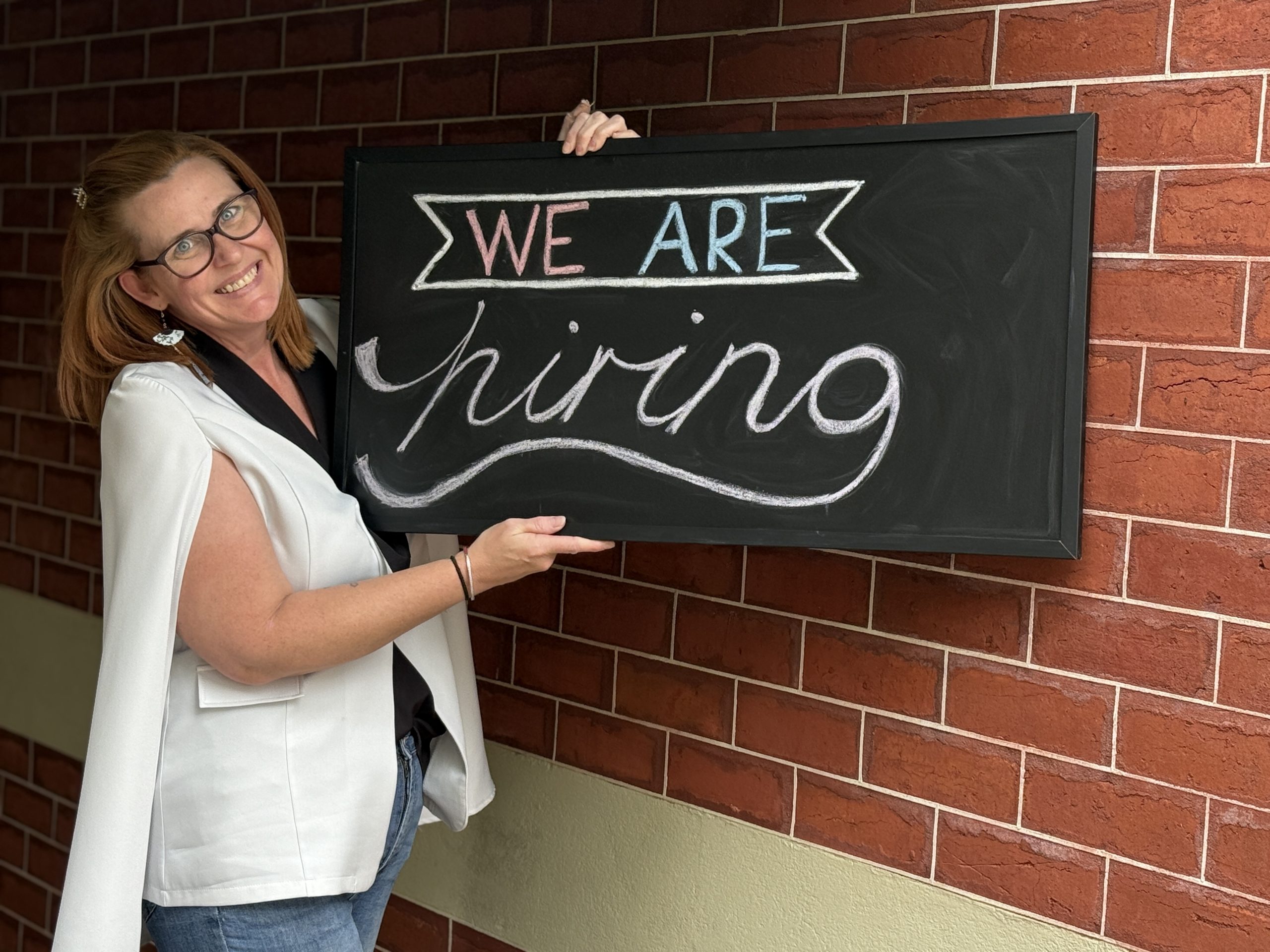 Woman holding 'We Are Hiring' sign against wall.
